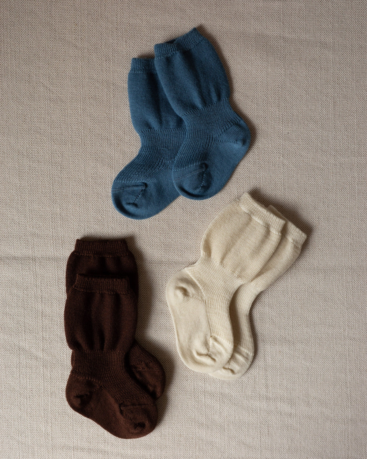Varig organic wool baby socks in blue, brown, and cream with ribbed cuffs, laid flat on a textured fabric surface.